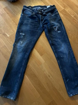 Blå jeans från Dondup, storlek 30 - Säljer ett par blå jeans från Dondup, tillverkade i Italien. Jeansen har slitna detaljer och små hål på framsidan samt diskreta färgstänk på benen. Klassisk femficksmodell med knapp och dragkedja i midjan. Materialet är jeans med en skön passform.