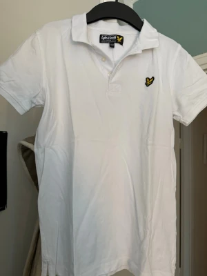 Vit piké från Lyle & Scott Junior - Säljer en klassisk vit pikétröja från Lyle & Scott Junior med korta ärmar och krage. Tröjan har två knappar vid halsen och en broderad gul fågel-logga på bröstet. Materialet är mjuk bomull som känns skönt mot huden. Perfekt för en clean och stilren look.