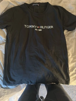 Svart t-shirt Tommy Hilfiger - Svart t-shirt från Tommy Hilfiger med broderad logga och texten 'EST. 1985' på bröstet. Klassisk rund hals och korta ärmar. Materialet känns mjukt och skönt, perfekt för en chill och stilren look.