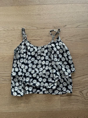 Svart blommig linnetopp med prästkragar - Säljer en svart linnetopp med vita och gula prästkragar över hela tyget. Toppen har smala axelband och ärmlös design, perfekt för varma dagar. Materialet känns lätt och luftigt, och mönstret är riktigt somrigt och fräscht.