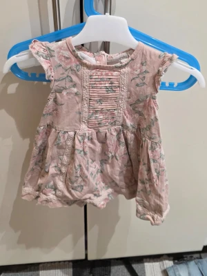 Blommig kortklänning från Newbie - Supersöt kortklänning från Newbie i storlek 80. Klänningen är ljusrosa med blommigt mönster och har fina spetsdetaljer samt veckad front. Tillverkad i mjuk bomull, perfekt för varma dagar. Korta ärmar och rund hals ger en gullig look.