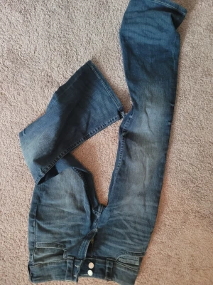 Blå bootcut jeans från H&M, barn - Säljer ett par blå bootcut jeans från H&M med låg midja och justerbar midja. Jeansen har snygga fickor med lock och silverfärgade knappar bak. Materialet är klassisk jeans med stretch med utsvängda ben nertill.