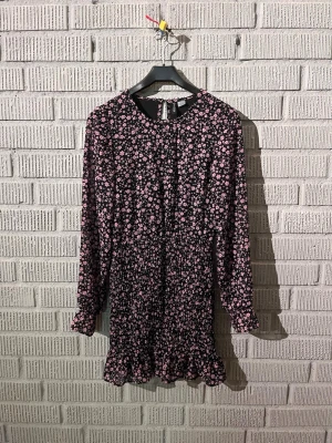 Svart blommig kortklänning H&M Divided - Supersöt svart kortklänning från H&M Divided med rosa blommigt mönster. Klänningen har långärmade ärmar, smockad midja och volangkant nedtill. Materialet är lätt och tunt, så inte särskilt varmt. Perfekt till fest eller kalas!