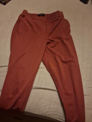 Rostbruna byxor från Vero Moda - Snygga rostbruna byxor från Vero Moda i rak modell med uppvikta benslut och resår i midjan. Byxorna har en enkel och clean look, perfekta till en chill vardagsoutfit. Materialet känns mjukt och skönt mot huden. Priset som står gäller, diskuteras helst inte🫶
