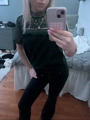 Mörkgrön spetsblus med volang - Säljer en mörkgrön blus i spets med korta ärmar och hög hals. Blusen har fina volangdetaljer och är genombruten med ett snyggt mönster. Perfekt att styla med smycken för en trendig look.