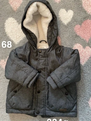 Mörkgrå vinterjacka från Newbie - Mysig mörkgrå vinterjacka från Newbie med huva och fluffigt vitt foder. Jackan har knappar framtill, två fickor och är perfekt för kalla dagar. Snygg och enkel design som håller värmen hela vintern.🌸 Fint använt skick. Storlek 68