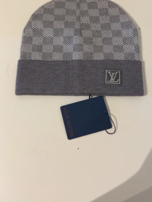 Grå rutig mössa - Snygg grå mössa från med rutigt mönster och uppvikt kant. Mössan har en broderad logga framtil och är stickad i mjukt material. Perfekt för dig som vill ha en lyxig accessoar med stilren design.