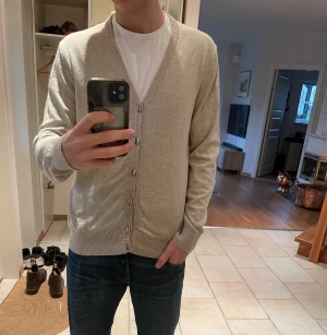 Stickad kofta från Stenströms - Säljer en stilren beige kofta från Stenström med unika, randiga knappar i flera färger. Koftan har v-ringning, ribbade muddar och är långärmad. Tyg från Zegna som det står på lappen i koftan och merinoull