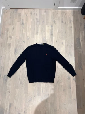 Mörkblå stickad tröja Polo Ralph Lauren - Klassisk mörkblå stickad tröja från Polo Ralph Lauren med rund halsringning och den ikoniska röda logon broderad på bröstet. Tröjan har långa ärmar och ribbade muddar vid ärmslut och nederkant. Perfekt för dig som gillar stilren och tidlös design.