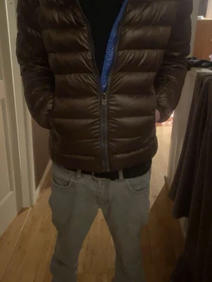 Brun Colmar pufferjacka med luva - Snygg brun pufferjacka från Colmar med blått foder i luvan och dragkedja framtill. Jackan har en rund Colmar-logga på ärmen, quiltad design och huva med tryckknappar. Perfekt för kalla dagar och har en modern look.