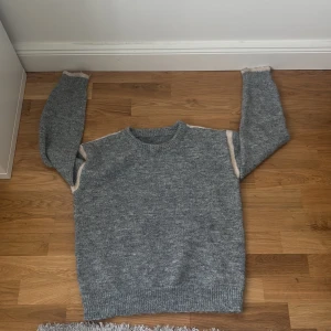 Grå stickad tröja med beige detaljer - Mysig grå stickad tröja med rund hals och långa ärmar. Tröjan har subtila beige detaljer längs axlarna och ärmarna, samt ribbstickade muddar vid hals, ärmslut och nederkant. 