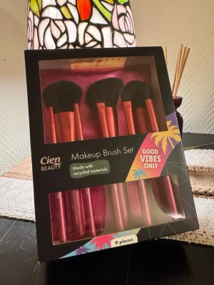 Cien Beauty Makeup Brush Set 9-pack - Säljer ett set med 9 sminkborstar från Cien Beauty. Borstarna har metallicrosa skaft och svarta borst, och är tillverkade av återvunna material. Perfekt för att applicera allt från foundation till ögonskugga. Kommer i en snygg förpackning med tropiskt tema.
