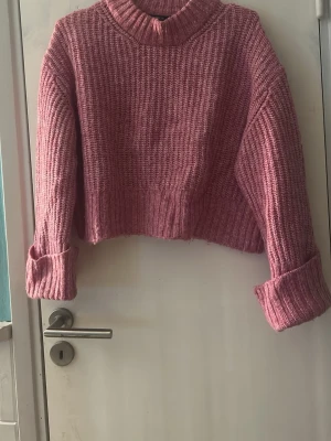 Rosa croppad stickad tröja - Säljer en mysig rosa stickad tröja med croppad passform och breda uppvikta ärmar. Tröjan har en hög krage och grov ribbad struktur som ger en cool och avslappnad vibe. Perfekt att matcha med jeans eller kjol för en trendig look.