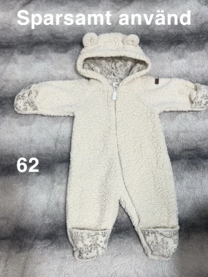 Vit fleeceoverall från Newbie - Supermjuk och gosig vit fleeceoverall från Newbie med öron på huvan och dragkedja framtill. Insidan har ett fint naturinspirerat mönster och händer samt fötter kan vikas över. Perfekt för kyliga dagar!🌸Sparsamt använd. Något plattare ”teddytyg” som det blir direkt. Övervikbara muddar, storlek 62