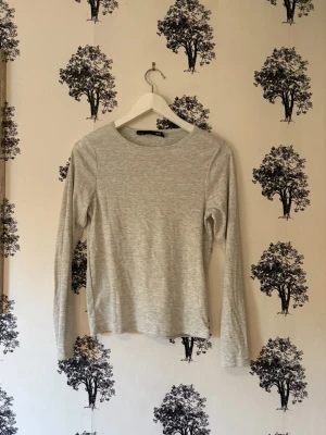 Ljusgrå långärmad topp  - En enkel och stilren ljusgrå långärmad topp från Lager 157. Toppen har rund halsringning och är gjord i mjuk bomullsmix med diskret ribbad struktur. Perfekt att matcha med jeans eller kjol för en clean look.