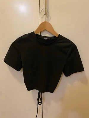 Svart topp med öppen rygg - Svart croppad topp korta ärmar och rund hals. Toppen har en stor öppen rygg med snörning som knyts baktill, strl S.