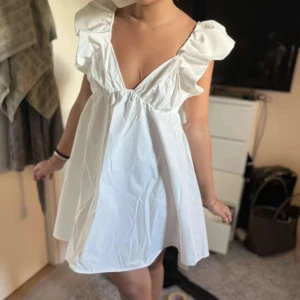 Vit volangklänning med rosett - Supersöt vit klänning med volangdetaljer vid axlarna och djup v-ringning. Klänningen har en öppen rygg med stor rosett och är ärmlös. Perfekt för sommaren