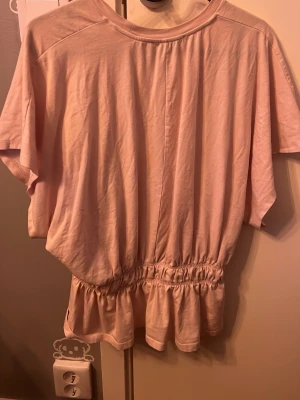 Ljusrosa blus med volang - Säljer en ljusrosa blus i mjukt bomullstyg. Toppen har korta ärmar, rund halsringning och en bred resår i midjan som ger en snygg volangkant nedtill. Perfekt för dig som gillar en feminin och avslappnad stil.