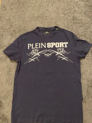 Mörkblå Plein Sport t-shirt - Snygg mörkblå t-shirt från Plein Sport med stort vitt tryck framtill. Trycket har coola tigerhuvuden och grafiska detaljer som ger en sportig vibe. Klassisk rund hals och korta ärmar. 