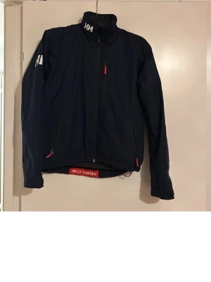 Mörkblå vindjacka från Helly Hansen - Snygg mörkblå vindjacka från Helly Hansen med HH-logga på kragen och axeln. Jackan har dragkedja framtill, två sidofickor med röda detaljer och hög krage. Perfekt för blåsiga dagar och har en clean, sportig look.