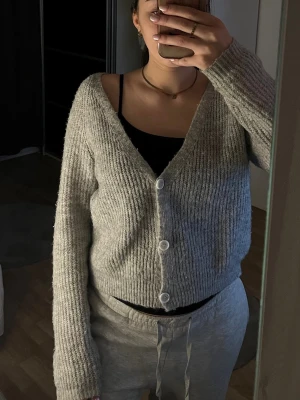 Grå ribbad kofta med knappar - Mysig grå kofta med ribbad struktur och tre vita knappar framtill. Koftan har en v-ringad hals och långa ärmar.