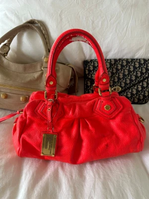 Orange handväska från Marc by Marc Jacobs - Så cool neonorange handväska i äkta läder från Marc by Marc Jacobs. Den ikoniska modellen Q Classic Baby Groove! En populär och eftertraktad vintagemodell med gulddetaljer och dubbla handtag. Långa bandet finns ej. Finns fläckar och slitage på lädret (se bild) men fortfarande en otroligt snygg vintage väska🤍  Mått: 34x18x12 cm (undersida) Kan hämtas hemma hos mig i Nacka eller skickas 🤍