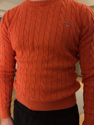 Orange kabelstickad tröja från GANT - Säljer en snygg orange kabelstickad tröja från GANT i premium bomull. Tröjan har rund hals, ribbade muddar och ett litet broderat GANT-logo på bröstet. Perfekt för dig som gillar klassisk stil med en färgklick. Storlek S.