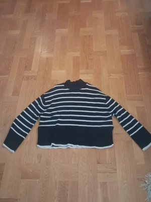 Mörk blå randig stickad tröja - Säljer en mörk blå stickad tröja med vita horisontella ränder och hög hals. Tröjan har lång ärm och en lite croppad passform med ribbad kant nedtill. Perfekt för dig som gillar klassisk stil med en modern twist. Den är använd max 5 gånger så det gör att den ungefär är i nyskick! 