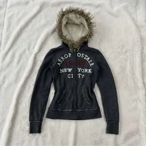 Aeropostale hoodie - Mörkgrå zip-hoodie från Aeropostale med tryck på bröstet: “Aeropostale 1987 New York City”. Hoodien har mjuk, fodrad huva med fuskpäls runt kanten samt två framfickor. Skön och varm modell som passar perfekt till vardags. Använd men i bra skick. (lånade bilder)