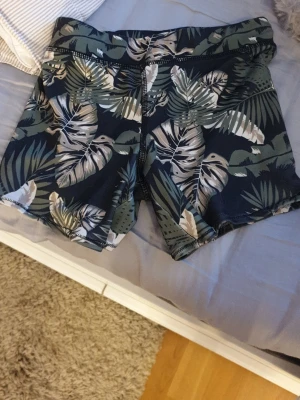 Mönstrade badshorts från Wave - Säljer ett par snygga badshorts från Wave med tropiskt bladmönster i grönt, beige och vitt mot en mörkblå bakgrund. Shortsen har snörning i midjan för perfekt passform och är gjorda i ett syntetmaterial som torkar snabbt. Perfekta för strand eller pool! Jag sydde de själv!