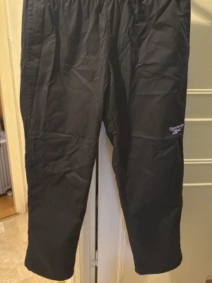 Svarta täckbyxor från Reebok - Svarta täckbyxor från Reebok med elastisk midja och diskret logga på benet. Byxorna har en enkel och sportig design, perfekta för utomhusaktiviteter. Materialet är syntet och ger ett lätt och smidigt intryck. Passar dig som gillar en clean och funktionell stil.