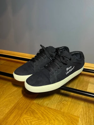 Saint Laurent sneakers - Svarta sneakers från Saint Laurent med vit logga på sidan och vit platt sula. Skorna har snörning och är tillverkade i canvas med detaljer i skinn. Tyvärr ingår inte box osv
