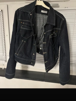 Dark Denim Cropped Jacket – Heine – Fitted Style - Stylish dark denim jacket from Heine with a flattering cropped fit. Features decorative stitching, zip details, and a unique patterned lining that adds a cool touch when worn open. Perfect for everyday wear or layering over dresses and tops. Brand: Heine Color: Dark blue / black denim Style: Cropped, fitted Details: Zip pockets, contrast stitching, patterned lining Condition: Very good condition (no visible flaws) Great for casual outfits, spring/autumn wear, or a chic street-style look. 📏 Pleas