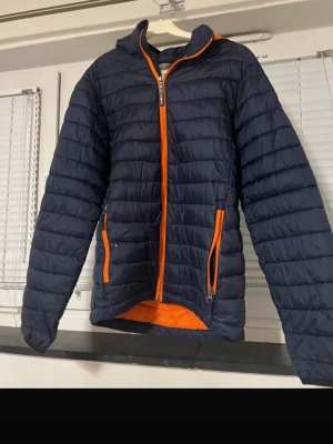 McKinley Kids Puffer Jacket - Lightweight and warm McKinley padded jacket for kids. Dark navy blue with bright orange details (zipper, pockets and lining). • Brand: McKinley • Size: 160 (fits approx. 10–11 years) • Color: Navy blue / Orange • Type: Puffer / insulated jacket • Closure: Full zip • Pockets: Two zip pockets ⚠️ Condition note: The jacket has a few small holes on the front (see close-up photos). They are minor and do not affect warmth, but are visible up close. Price reflects this. Perfect as a spare jacket for sch
