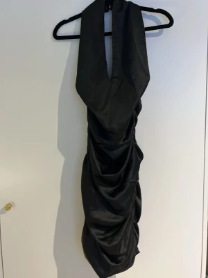 Svart långklänning i satin - Säljer en svart långklänning med djup v-ringning och draperad effekt längs hela klänningen. Klänningen har en snygg omlottkänsla upptill, dragkedja bak och är öppen i ryggen. Materialet har en glansig satinfinish som ger en lyxig känsla. Storlek 36 Tall så perfekt om man är lång.