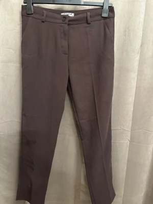 Bruna kostymbyxor från Esila - Snygga bruna kostymbyxor från Esila med klassisk rak passform och midwaist. Byxorna har bälteshällor, knapp och dragkedja framtill samt diskreta sidofickor. Materialet är mjukt och känns som bomull, perfekt för en clean och stilren look.