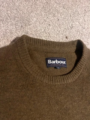 Olivgrön stickad tröja från Barbour - Säljer en olivgrön stickad tröja från Barbour med rund hals och ribbade muddar. Perfekt för dig som gillar klassisk stil och vill ha något som funkar till både jeans och chinos. Materialet är mjukt och värmande, och färgen är diskret men snygg.