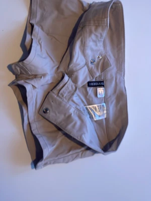 Beige shorts från Divided - Säljer ett par beige shorts från Divided med klassisk design och fickor med knappdetaljer. Perfekta för varma dagar och har en avslappnad stretchig passform. Materialet är mjukt och lätt  som känns skönt mot huden.