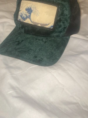 Grön/svart Velvet keps med Våg motiv 🌊  - Säljer en unik truckerkeps från Hatstore i mörkgrön sammet med svart mesh baktill. Framtill finns en beige patch med blått vågmotiv. Kepsen har justerbar snapback och cool vintagekänsla. Perfekt för dig som vill sticka ut med din stil.
