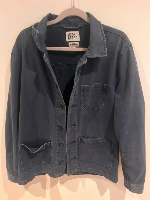 Blå overshirt från Roots - Säljer en blå overshirt från Roots med klassisk krage och tre stora fickor framtill. Jackan har knappar hela vägen och är tillverkad i ett mjukt bomullsmaterial. Perfekt för lager på lager och enkel att styla.