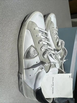 Philippe Model vita sneakers - Snygga vita Philippe Model sneakers med grå mockadetaljer, svart häl och en metallic-silver patch på sidan. Skorna har vita snören med svart text och en platt sula. Tillverkade i skinn och mocka, perfekt för dig som gillar stilrena men ändå unika sneakers.