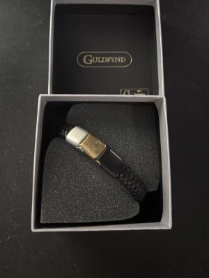 Armband - Säljer ett svart flätat armband från Guldfynd med detaljer i silver och guld. Har aldrig använt de. Längden på armbandet är 21 cm.
