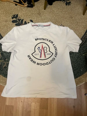 Vit Moncler t-shirt med tryck - Säljer en vit t-shirt från Moncler i storlek M med stort tryck på bröstet i svart, blått och rött. T-shirten har korta ärmar och rund hals med snygg detalj i form av röd och blå rand på insidan av kragen. Materialet är mjuk bomull och på ärmen finns en broderad Moncler-logga.