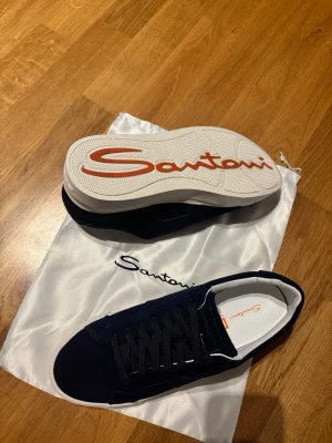 Mörkblå sneakers från Santoni - Snygga mörkblå sneakers från Santoni med vit platt sula och orange logga undertill. Skorna har snörning och är tillverkade i mocka med vita detaljer på insidan. Perfekta för dig som gillar stilrena och exklusiva sneakers.