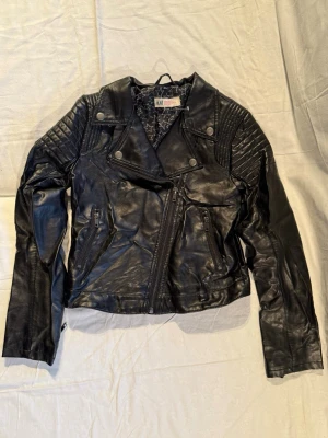 Svart skinnjacka från H&M - Cool svart skinnjacka från H&M med quiltade detaljer på axlarna och dragkedjor framtill. Jackan har klassisk bikerstil med snedställd dragkedja och långa ärmar. Fodret har ett snyggt stjärnmönster som ger extra attityd.