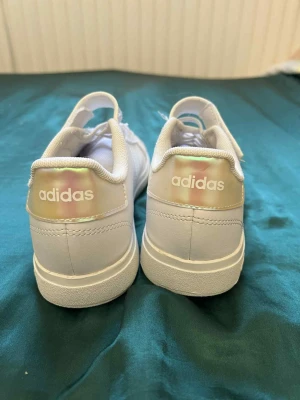 Vita adidas sneakers med holografiska detaljer - Snygga vita sneakers från adidas med holografiska ränder och häl. Skorna har snörning och kardborreband för extra stabilitet. Ovandelen är i syntetmaterial och sulan har ett coolt hexagonmönster. Perfekt för dig som gillar stilrena och moderna sneakers.
