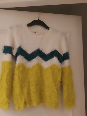 Gul och vit fluffig stickad tröja H&M storlek 134/140 - Fluffig stickad tröja från H&M i storlek 134/140. Tröjan har breda zigzag-ränder i mörkblått, vitt och gult, samt långärmad modell. Materialet är mjukt och lurvigt, perfekt för att hålla sig varm och se cool ut under hösten och vintern.