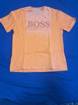 Orange t-shirt från Hugo Boss - Snygg orange t-shirt från Hugo Boss med stort BOSS-tryck på bröstet. Klassisk rund hals och korta ärmar. Tillverkad i mjuk bomull som känns skön mot huden. Perfekt för dig som gillar stilrena logotoplagg och vill sticka ut med färg.
