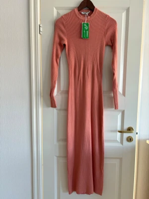 Rosa ribbad långklänning från NA-KD M - Snygg långklänning i en mjuk rosa ton från NA-KD. Klänningen är ribbad, har lång ärm och en rund halsringning. Materialet är stickad och stretchigt vilket ger en slim passform. Perfekt för dig som gillar enkel och stilren design.