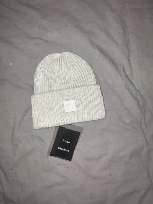 Acne Mössa - Bilderna visar en vit stickad mössa från Acne Studios med uppvikt kant och deras ikoniska ansiktslogga framtill. Ingen väst syns på bilderna.